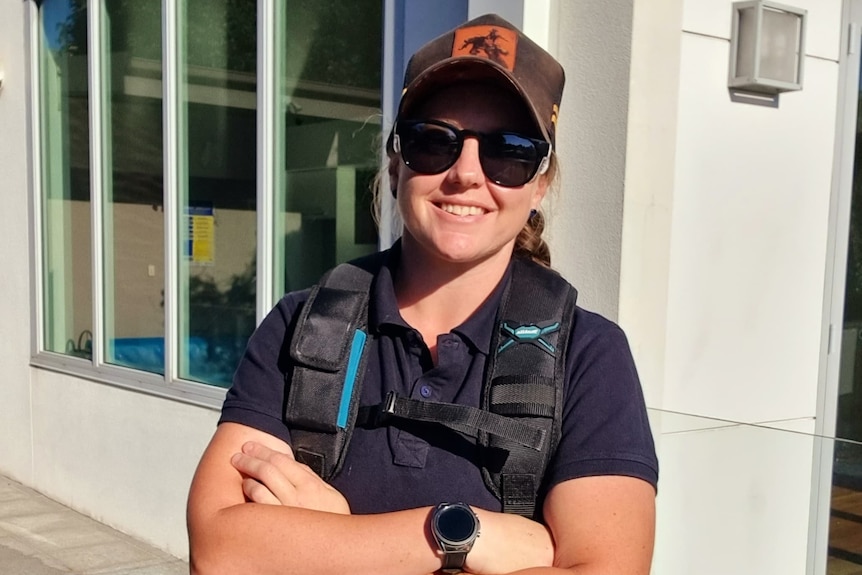 Une jeune femme souriante porte un capm lunettes noires, un gilet de protection et un t-shirt violet, se tient les bras croisés.