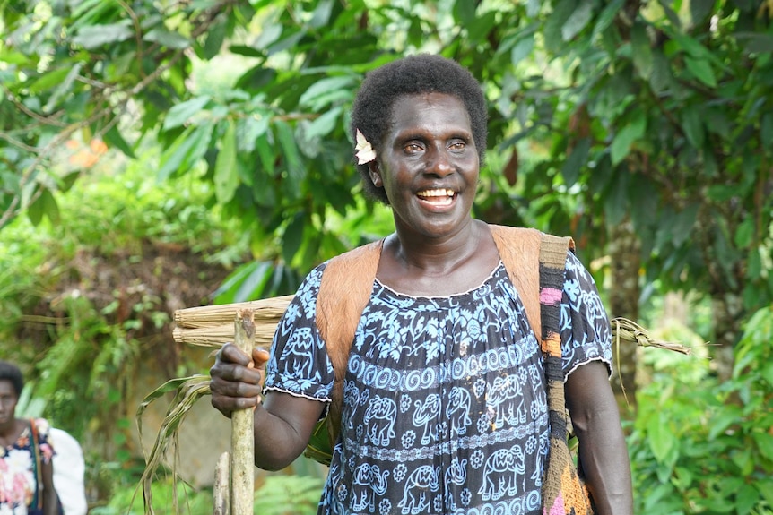 Susan Paai smiles as she holds a stick with a basket full of vegetables on her back.