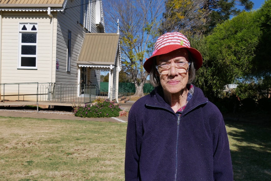 An elderly lady wearing a purple jacket and hat standing outside a church. 
