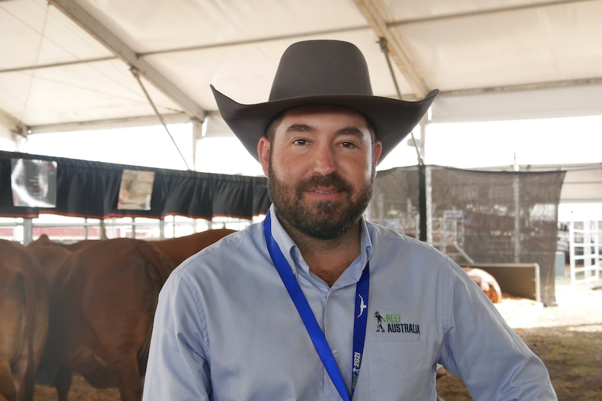 A man with brown hair wearing an akubra. There are cows in the background.