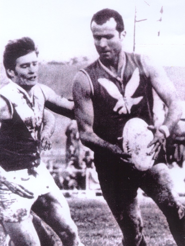 A SANFL footballer holds the ball, preparing to kick while an opponent tries to close in. 