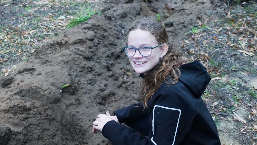 A young girl in the forest smiling while planting a tuart seedling.