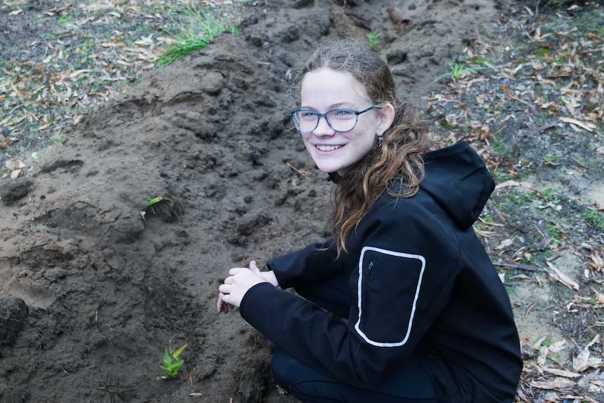 A young girl in the forest smiling while planting a tuart seedling.