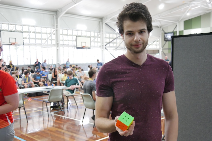 A man in maroon shirt holds a Rubik's Cube in a hall in front of an audience.