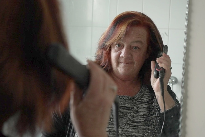 A woman in front of a bathroom mirror using a hair straightener.