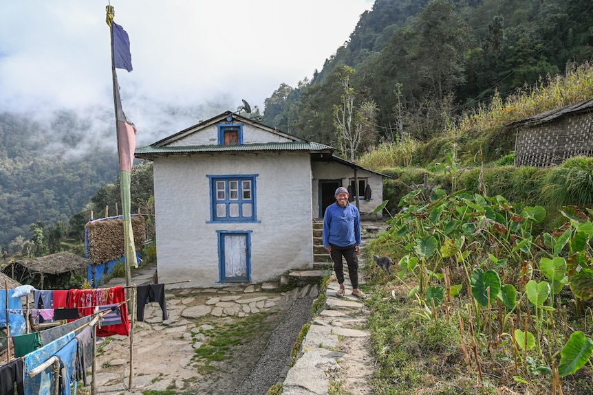 A man in a blue jumper stands outside a white house with blue painted windows and mist in a valley behind