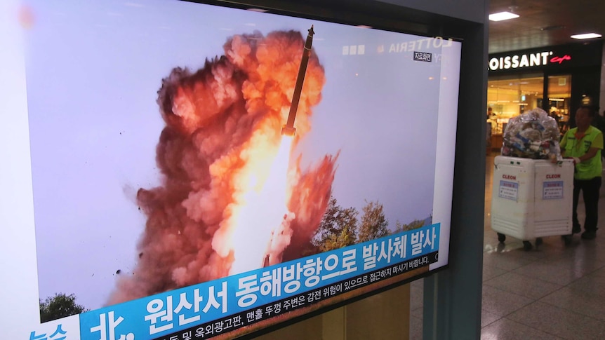 A man with a cleaning cart in a shopping watches television footage of a missile being launched.