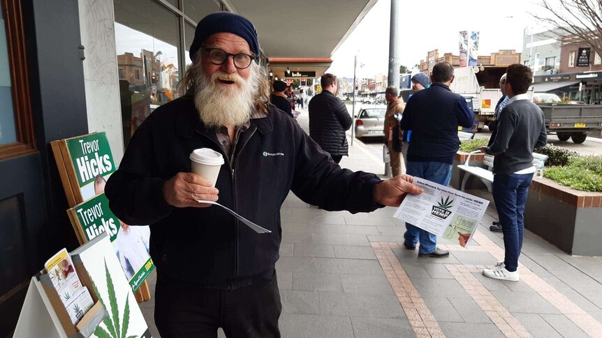 An elderly fellow with a voluminous white beard holding a coffee cup and handing out voting guides on the street.