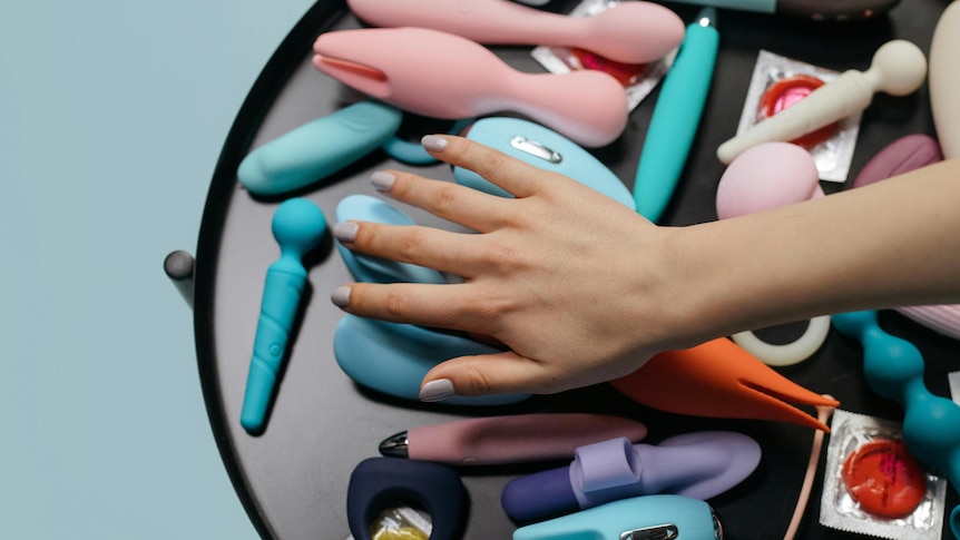 a hand hovering over a bunch of different coloured sex toys vibrators and condoms