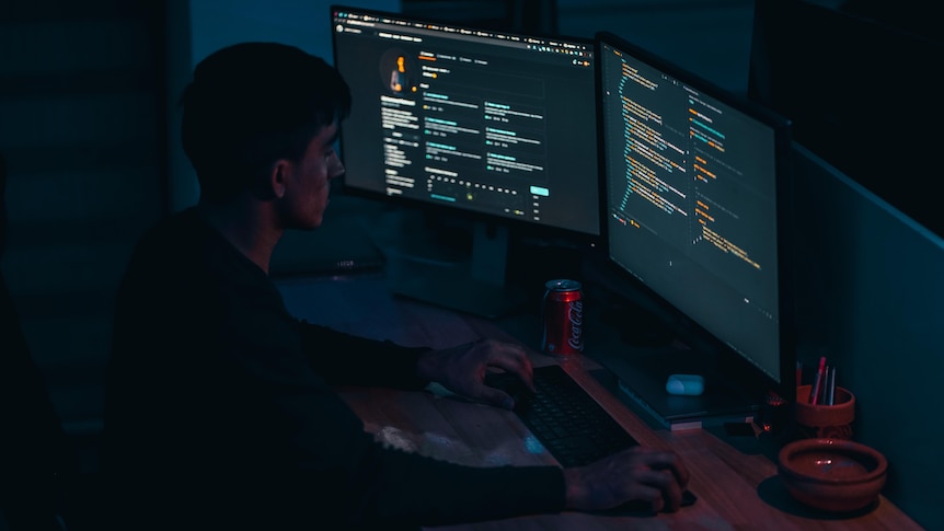 A man sitting in a dark room in front of multiple monitors showing code