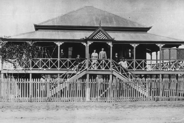 A black and white photo of a family standing on the top step of a house built on stilts, with wrap-around, covered  verandas.