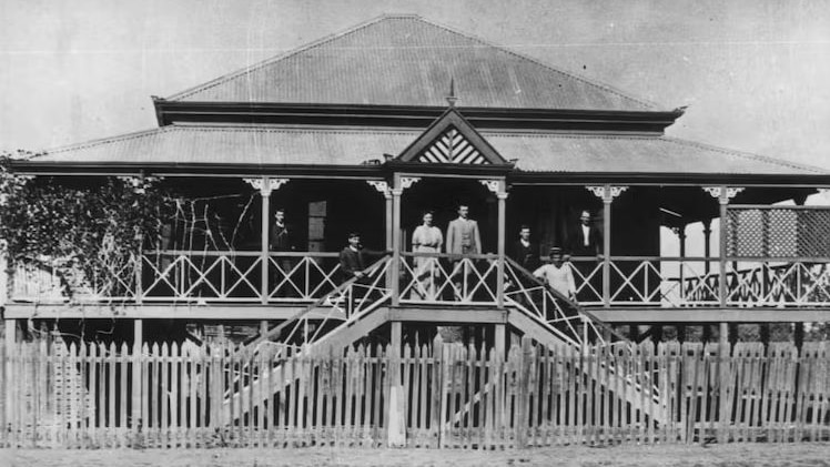 A black and white photo of a family standing on the top step of a house built on stilts, with wrap-around, covered  verandas.