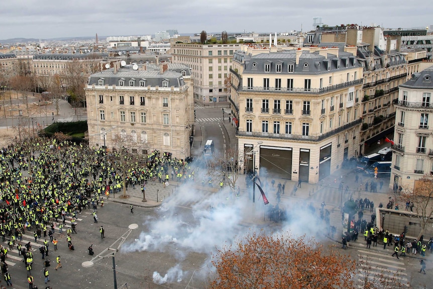 A Paris square is photographed from above. A crowd of protesters in yellow vests fill one side, while tear gas approaches.