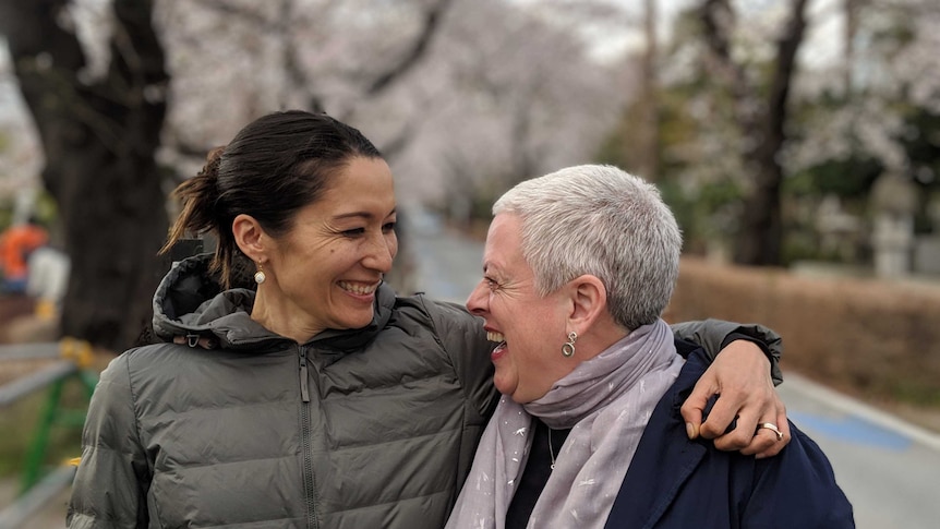 Two women embrace and smile under a canopy of cherry blossoms in Japan.