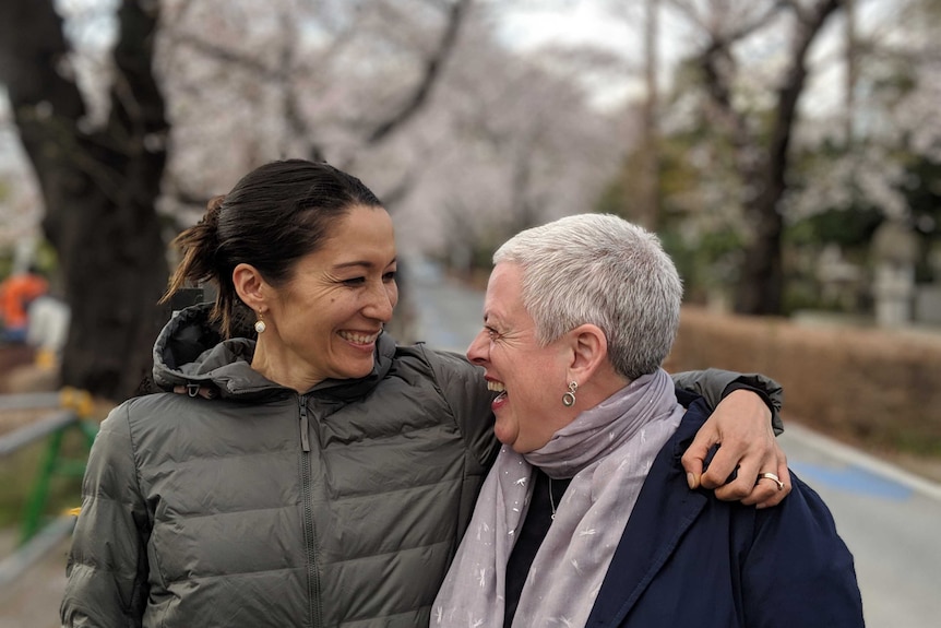 Two women embrace and smile under a canopy of cherry blossoms in Japan.