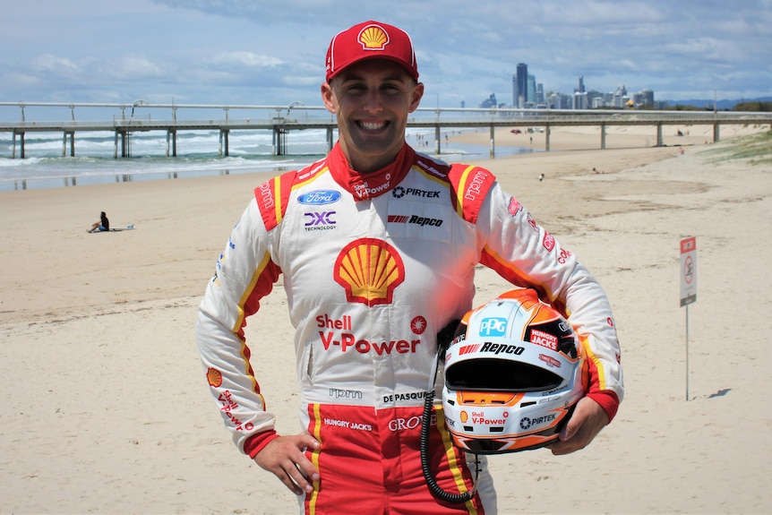 Man in race car suit standing on beach while holding a helmet. 
