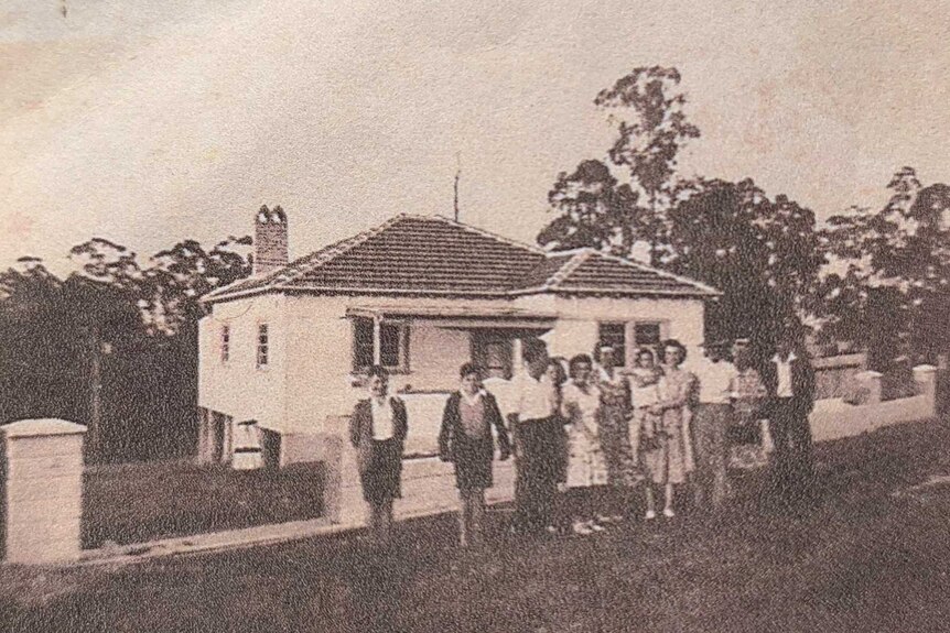 A black and white photo of a cottage in the 1950s with people standing in front of the building.