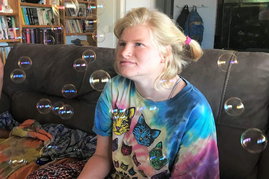 A young woman with blond hair, blue coloured eyes sits on a couch with bubbles all around her, wearing a bright tie dye shirt.