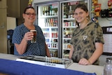 Two women smile at the camera while standing behind a bar, one holding a beer.