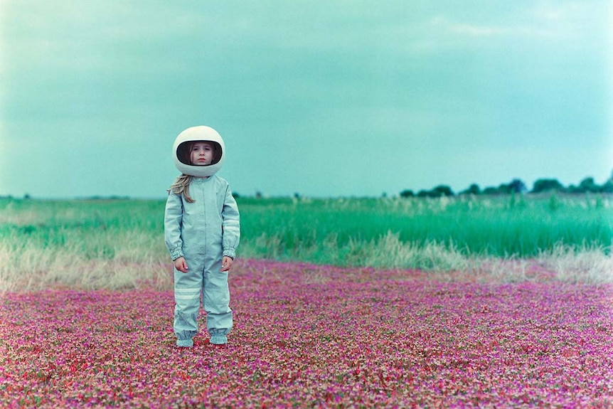 A young girl wearing a spacesuit costume and helmet stands in a field of purple flowers under an overcast sky