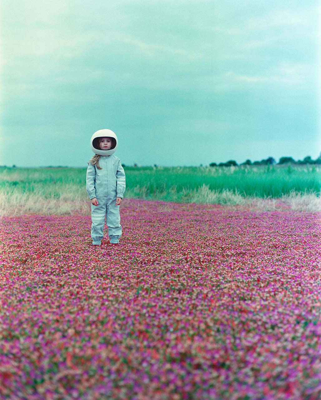 A young girl wearing a spacesuit costume and helmet stands in a field of purple flowers under an overcast sky