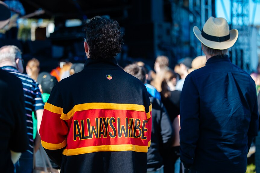 A man wearing a jacket that says "Always will be" stands among a festival crowd.