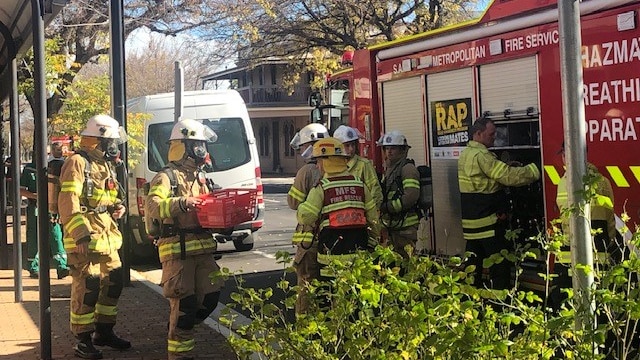 Seven firefighters stand alongside a fire truck on the footpath outside of a building