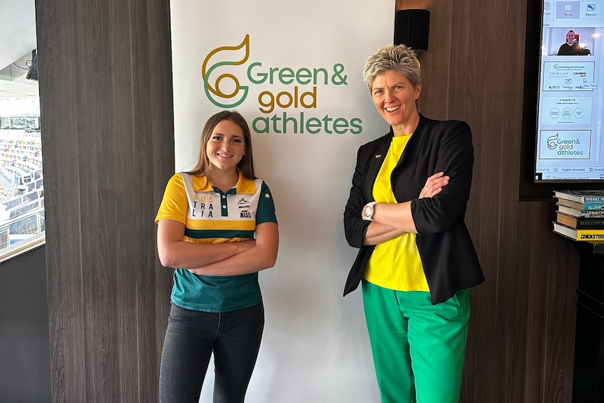 A woman and a teenage girl in green and gold smile with arms folded in front of a banner that says Green & Gold Athletes.