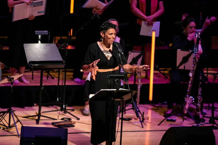 An Aboriginal woman dressed in black stands at a microphone with a slight smile in front of a classical ensemble on stage