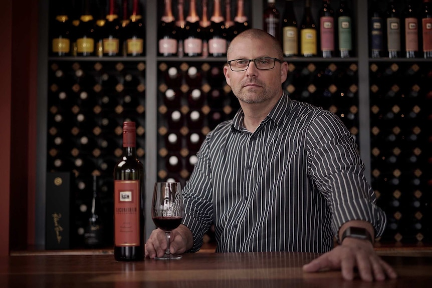 Damien White stands at a bar with a glass of wine in front of him and tens of bottles of wine on a shelf behind him.