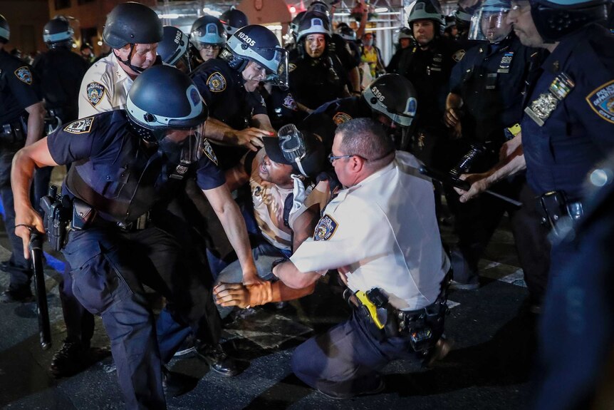 A black man is held by four white police officers wearing helmets in a night-time rally.