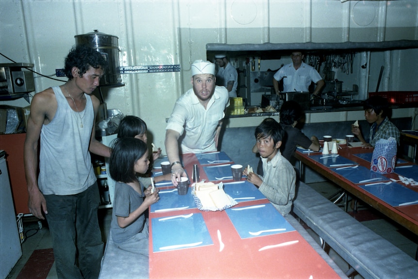 Australian Navy members feed Vietnamese children and adults at bench tables and seating aboard a warship