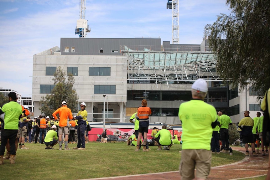 Workers in hi-vis clothing gathered on grass at Curtin University looking at the collapsed roof of a building.