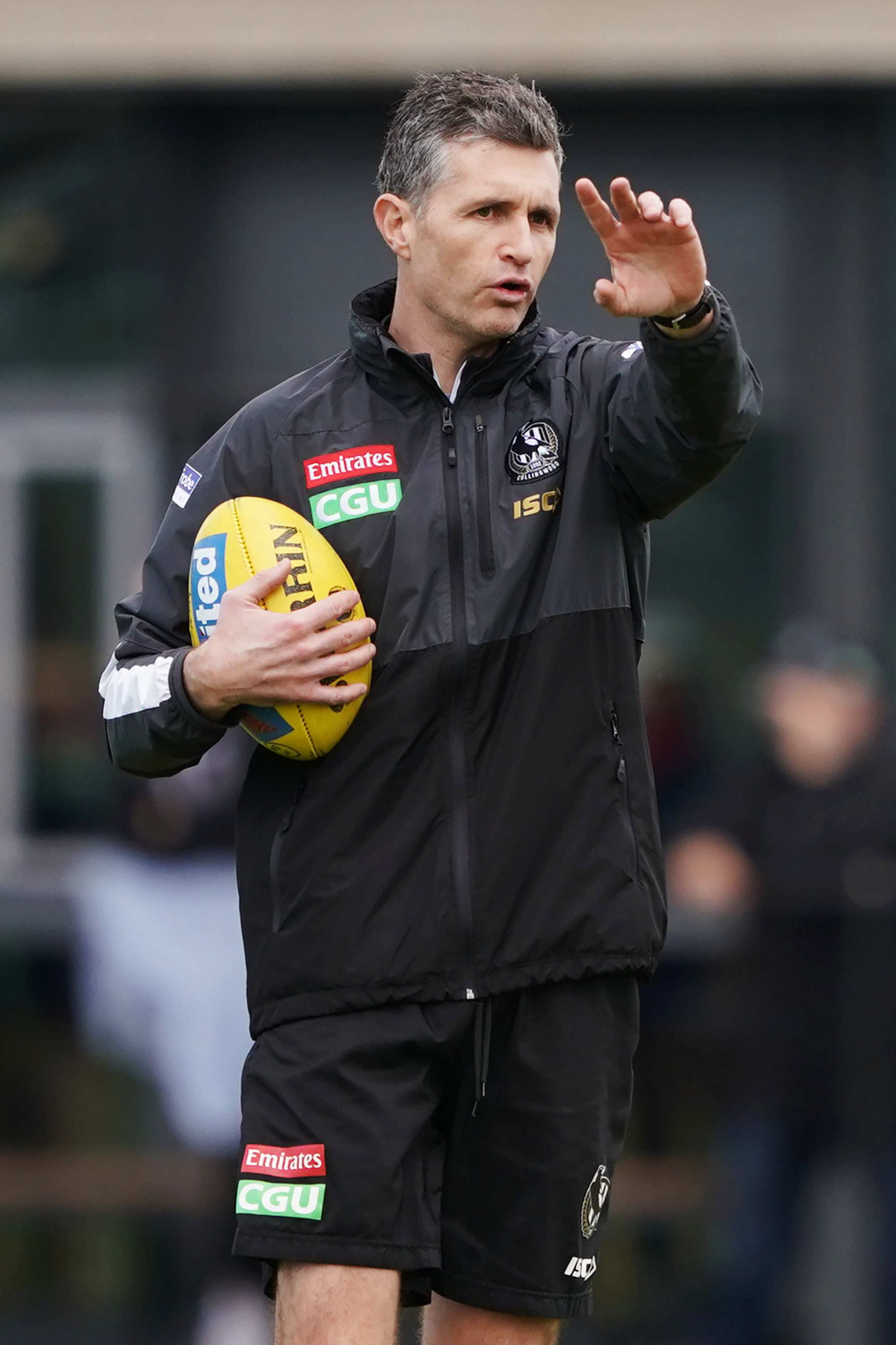 Justin Longmuir stands giving directions with one arm outstretched and the other holding a football during Collingwood training.