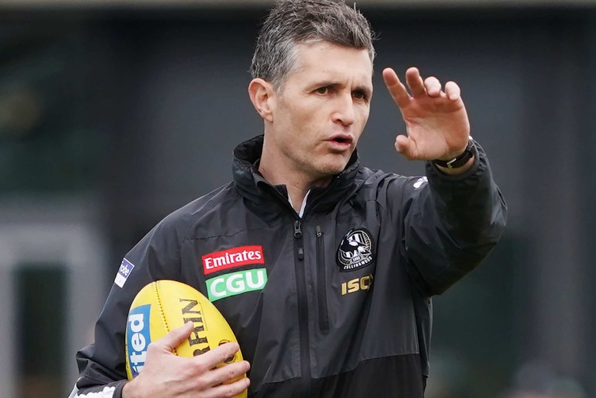 Justin Longmuir stands giving directions with one arm outstretched and the other holding a football during Collingwood training.