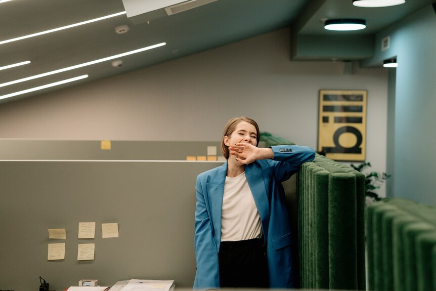 A woman in a blue jacket stands in an open plan office cubicle, yawning