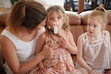 A woman sits on a couch helping a young girl with a breathing apparatus while another girl sits the other side of her