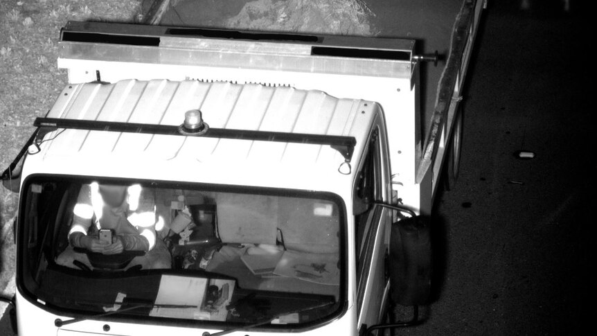 A black and white image of a person at the wheel of a dump truck with both hands on a mobile phone.