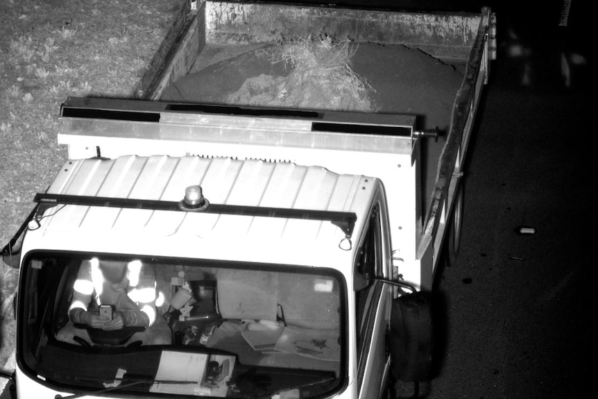 A black and white image of a person at the wheel of a dump truck with both hands on a mobile phone.