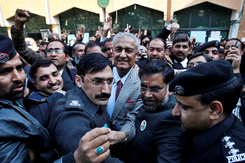 Smiling man in suit is surrounded by policemen and lawyers in a large crowd
