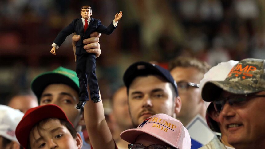 A boy holds a figurine of US President Donald Trump at his 100 days in office rally in a crowd of people wearing caps