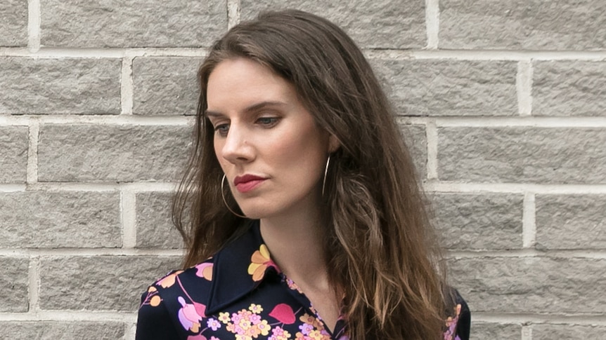 Leslie Jamison, with brown hair and flower print dress, stands against a grey wall looking down and to side with serious face.