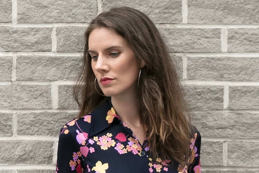 Leslie Jamison, with brown hair and flower print dress, stands against a grey wall looking down and to side with serious face.