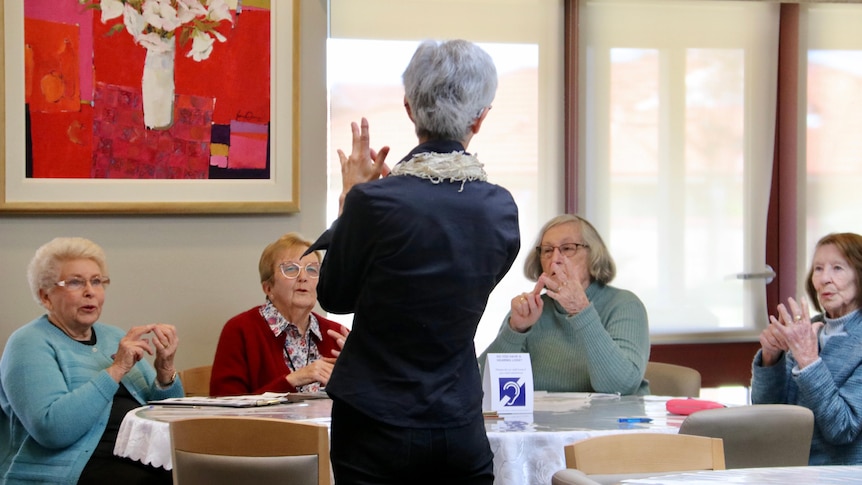 A woman in a black shirt with grey hair faces away from the camera doing a hand sign, with four elderly women in front of her. 