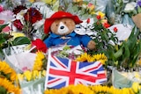 A Paddington bear toy sits in the centre of a pile of flower bouquets and UK flags. 