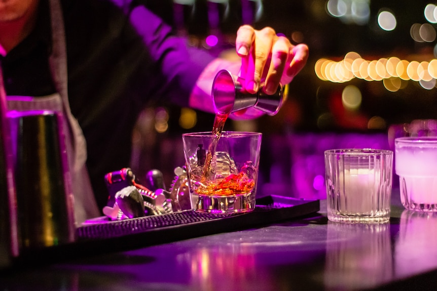 A bartender, face unseen, fixes a drink in a nightclub.