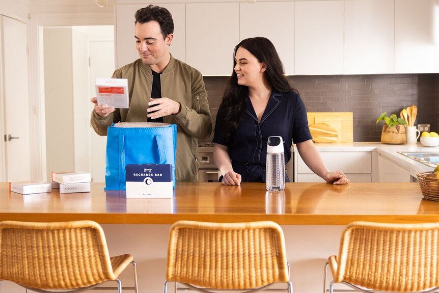 A man and a woman unpack a bag filled with cardboard containers in a bright kitchen.