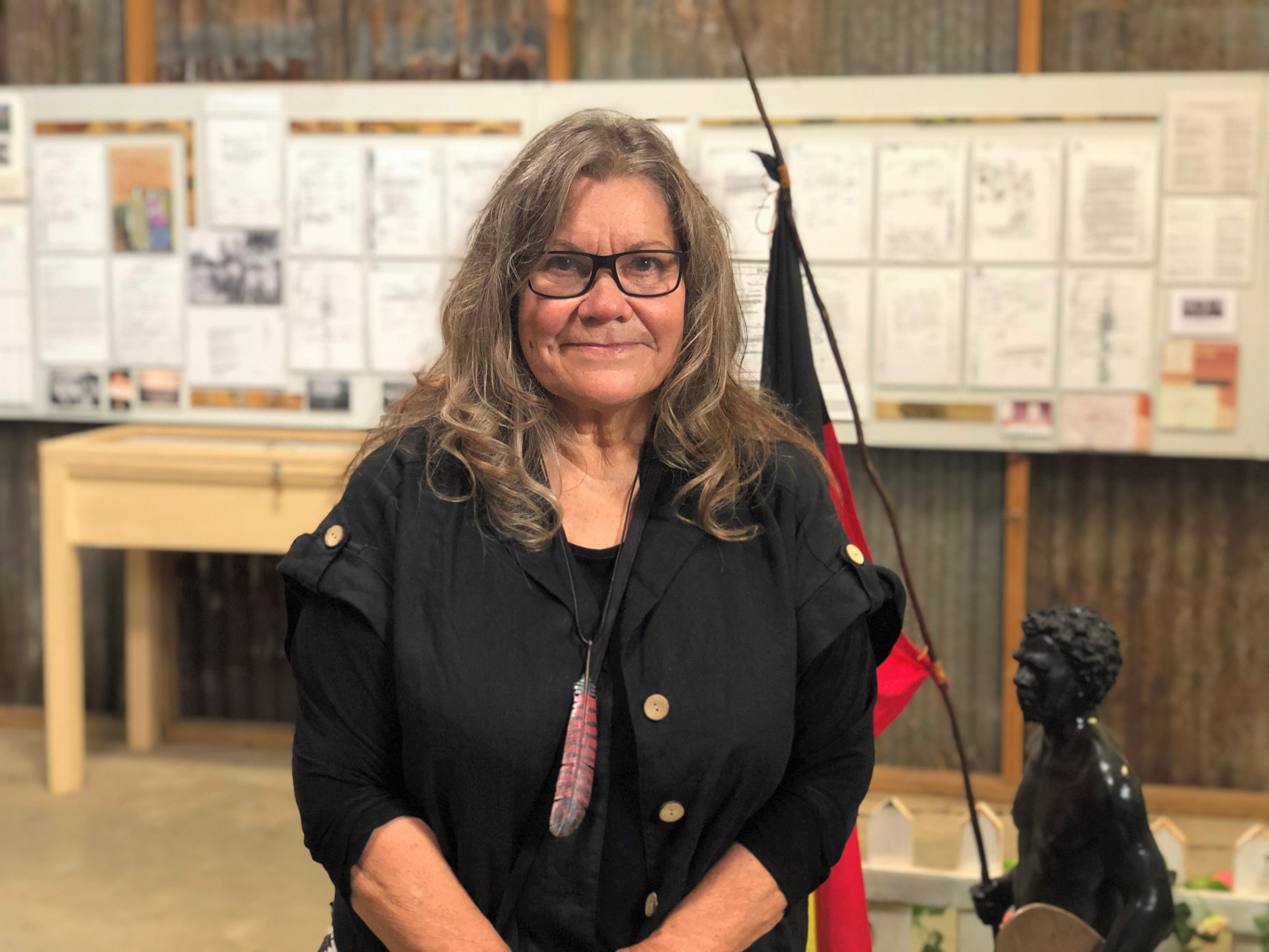 Woman smiles inside exhibition in front of statue with Aboriginal flag and historical documents on wall. 