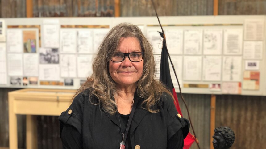 Woman smiles inside exhibition in front of statue with Aboriginal flag and historical documents on wall. 