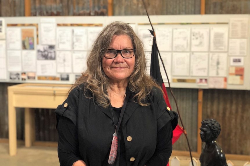 Woman smiles inside exhibition in front of statue with Aboriginal flag and historical documents on wall. 